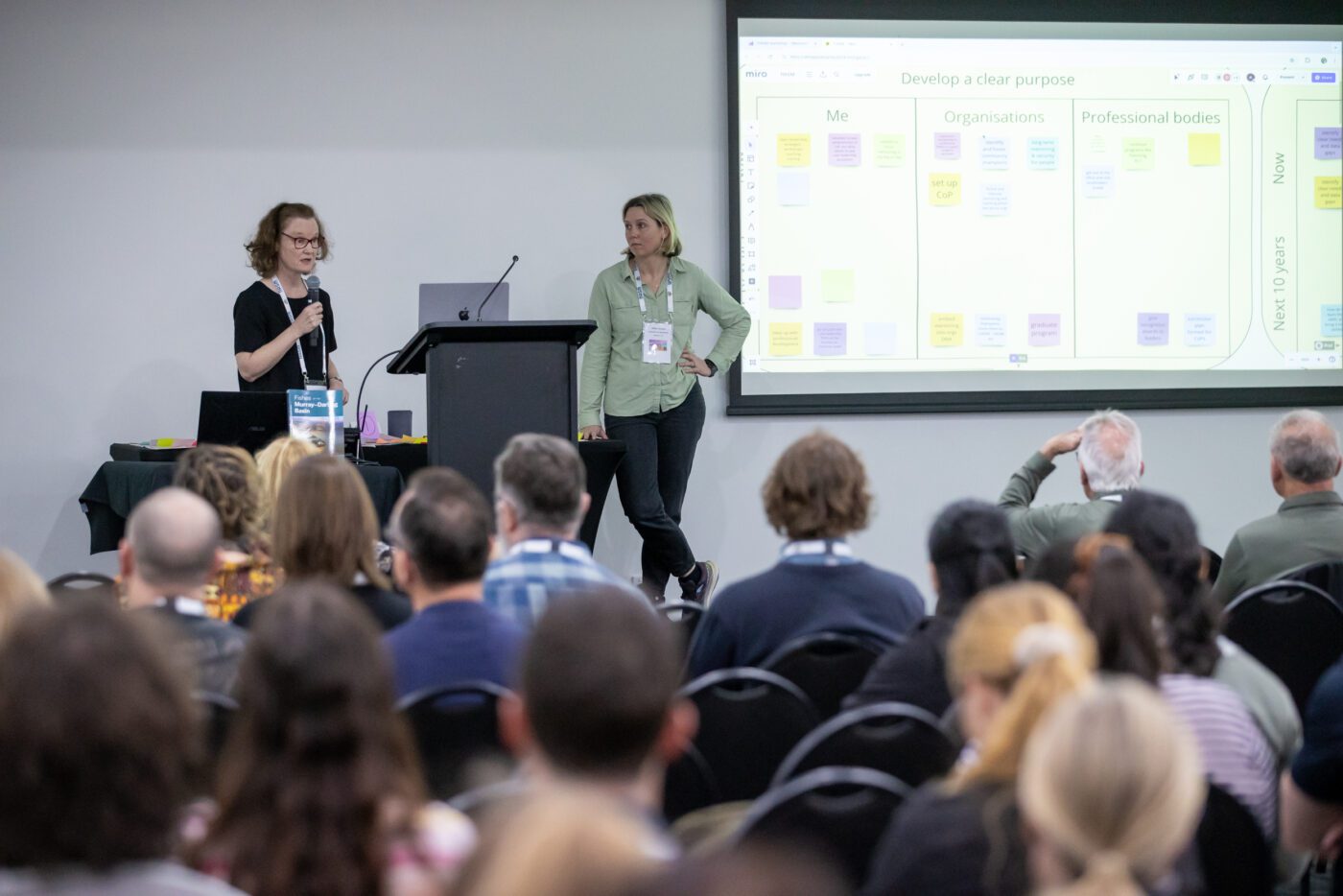 Two women on stage presenting in front of a seated crowd