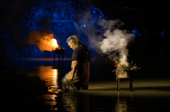 Man standing in water with smoking ceremonial fires in background